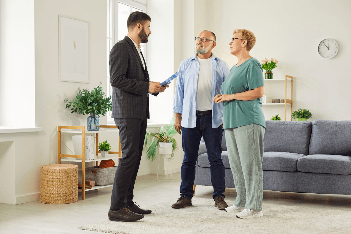 Male talks through home features with a mature senior couple during house visit, focusing on layout and living room appeal details.