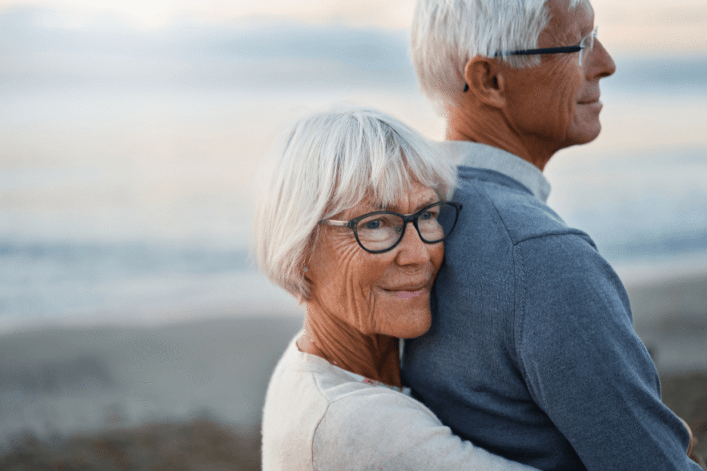 Senior woman holding her partner from behind on the seashore at sunset.