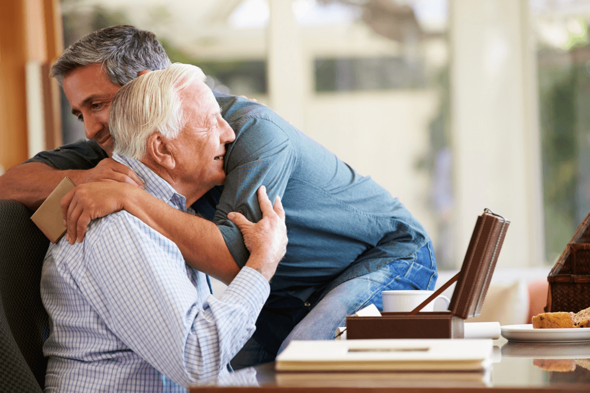 Two adults sharing a warm embrace in a bright room with a table holding a box, cup, and plate with food, reflecting a community lifestyle moment at Glen Carr.